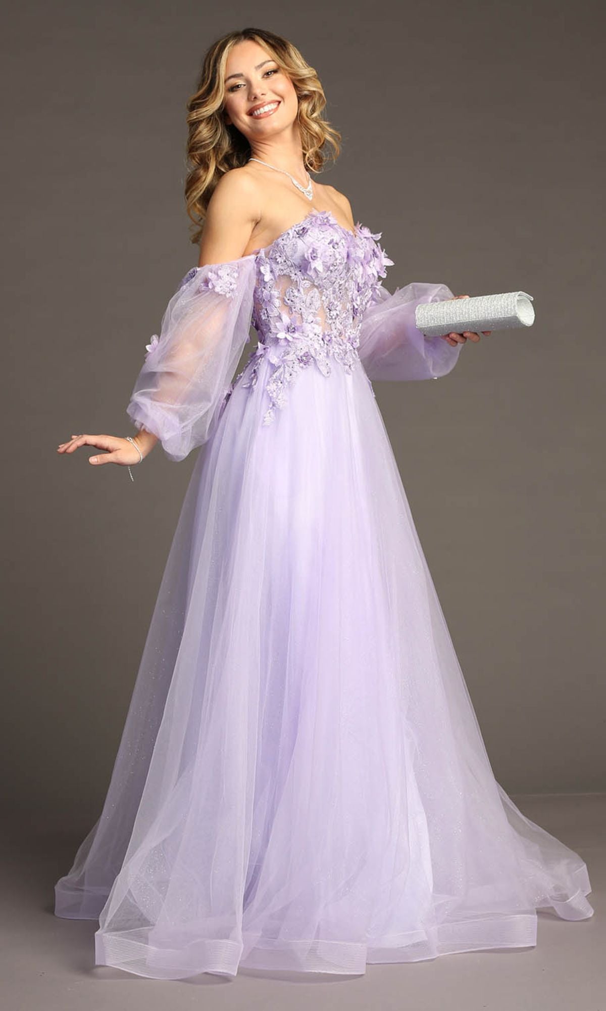 A woman wearing the Long Sleeve Long Prom Ball Gown Chicas CHF3096, featuring a light purple off-the-shoulder tulle skirt with floral details and holding a sparkling silver clutch, stands against a plain background.